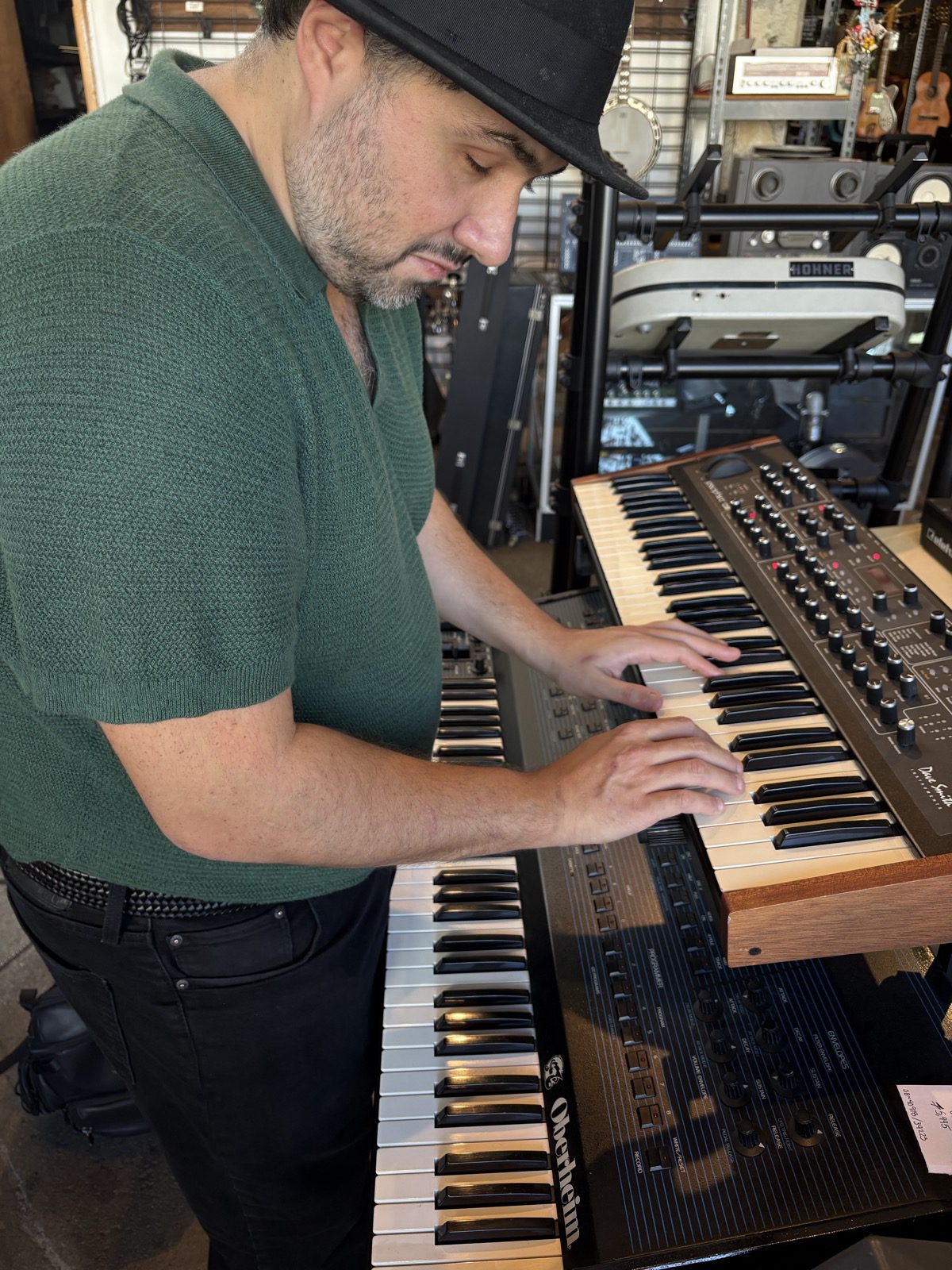 Sina standing at the Prophet in the shop, head bowed toward the keys, both hands on the keyboard, mid-chord. Sina wears a black hat, a green knit short-sleeved polo, and black jeans. The Prophet sits on top of a bigger black Oberheim keyboard. Behind, a white Hohner Clavinet on a stand, guitars and a banjo on the wall, and racks of gear.